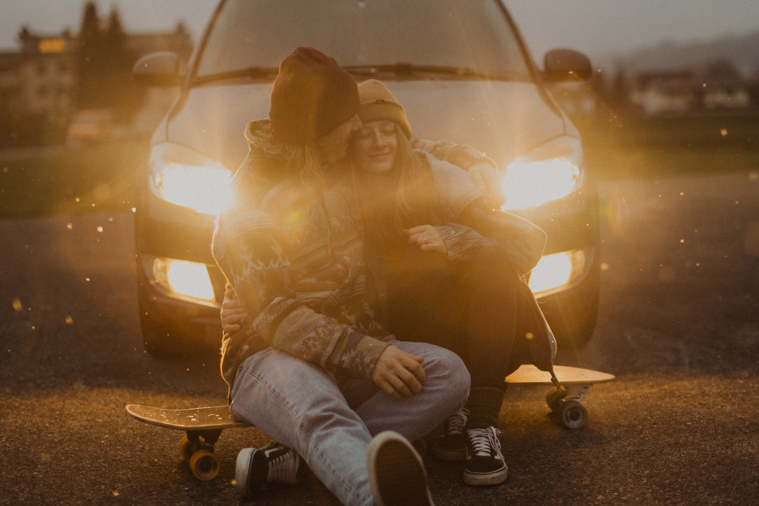 Paar sitzt auf Skateboard vor einem Auto in der Dämmerung. Es Schneit.