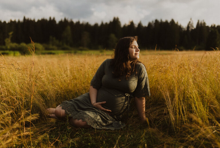 Frau sitzt in hohem Gras und hält den Babybauch. Das Gesicht zur Sonne, es ist Sommer und Sonnenuntergang.