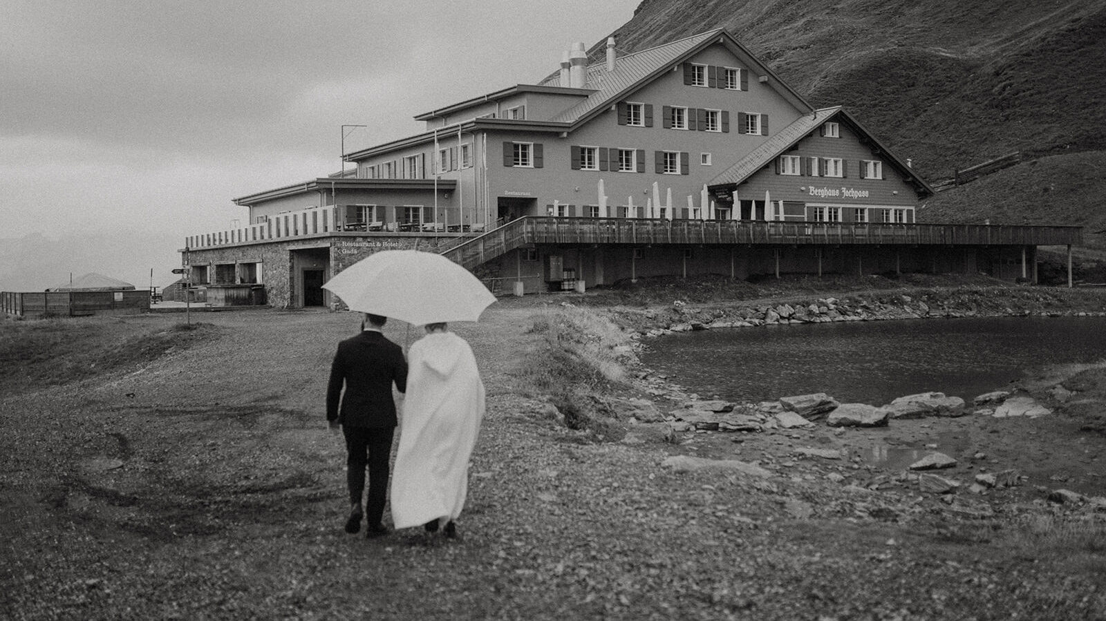 Berghochzeit auf dem Jochpass im Oktober. Brautpaar läuft unter einem Regenschirm auf das Berghaus Jochpass zu.