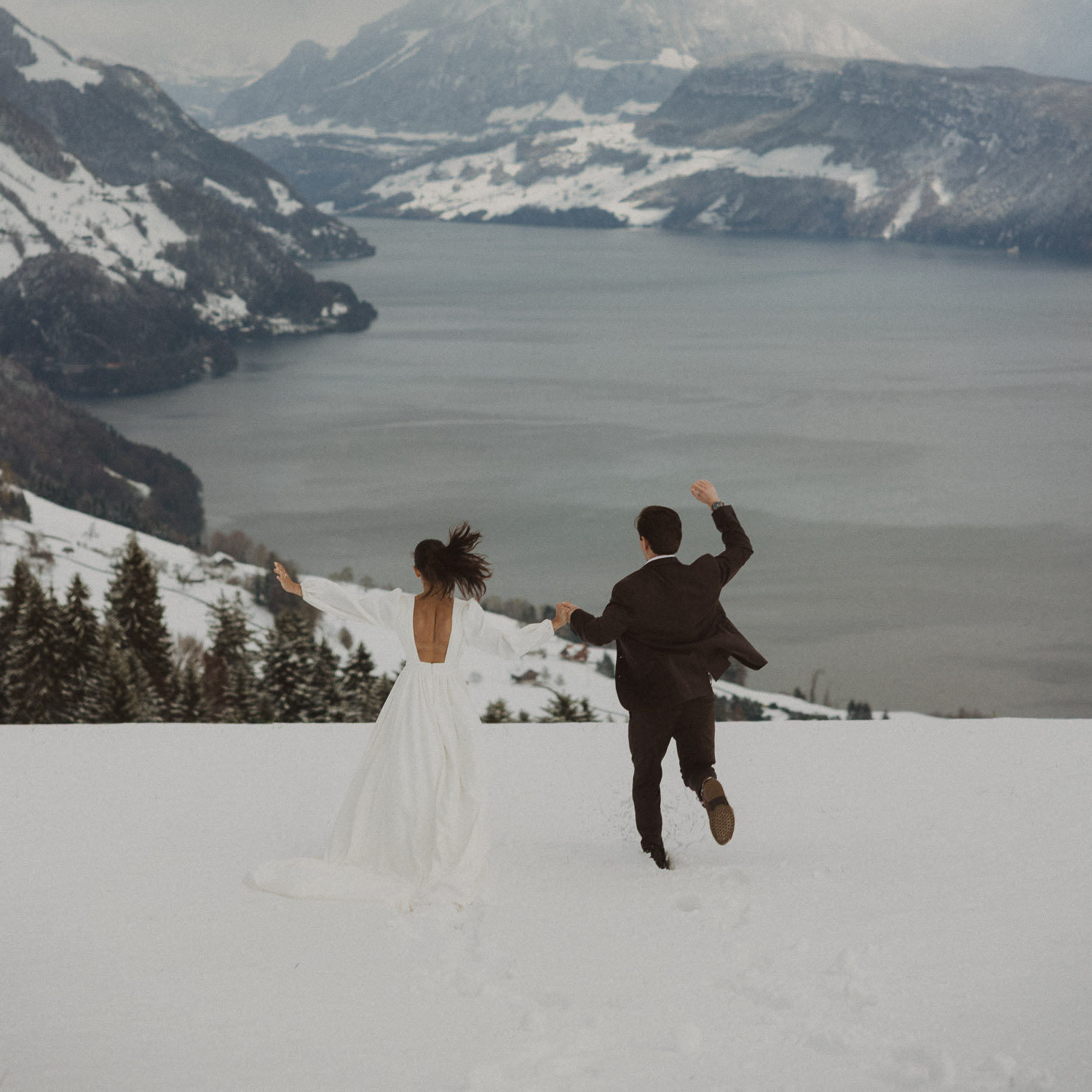 Couple running during an elopement photography session in Lucerne with snow-covered alpine mountains and a lakeside backdrop.