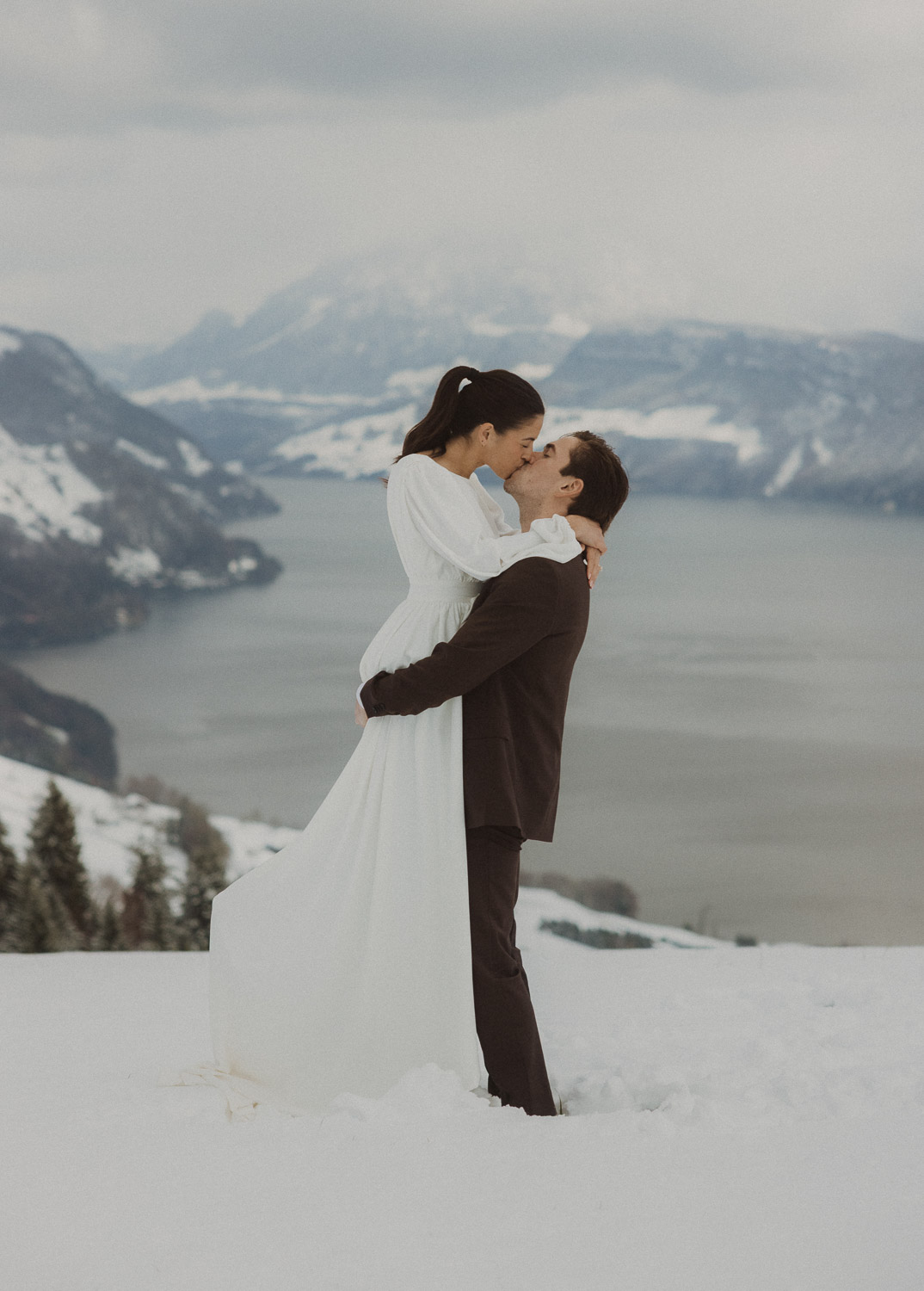 Couple kissing during an elopement photography session in Lucerne with snow-covered alpine mountains and a lakeside backdrop.